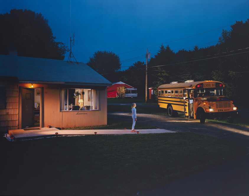 Child stands on driveway at dusk facing a waiting yellow school bus with door open.
