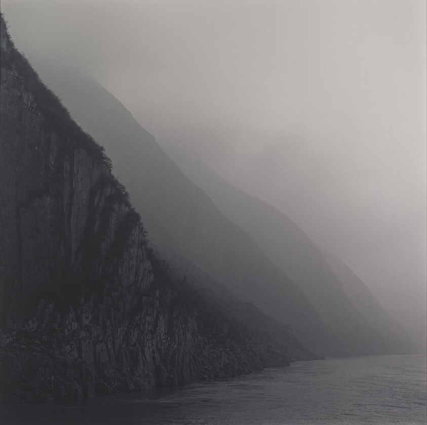 Steep rocky cliffs and layered mountain ridges fade into mist above a calm river.