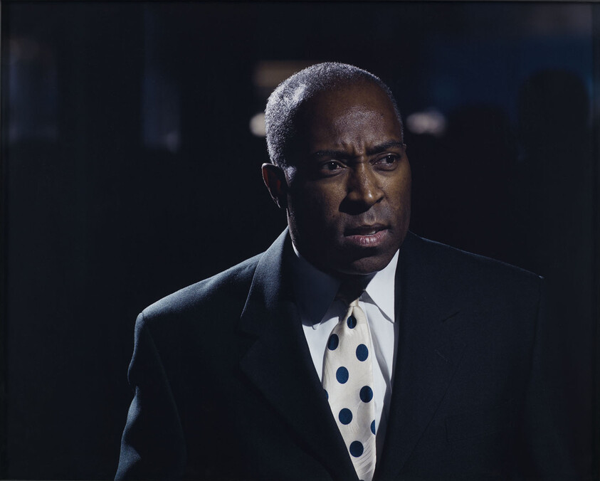 A man in a dark suit and white polka-dot tie looks serious in dim lighting.
