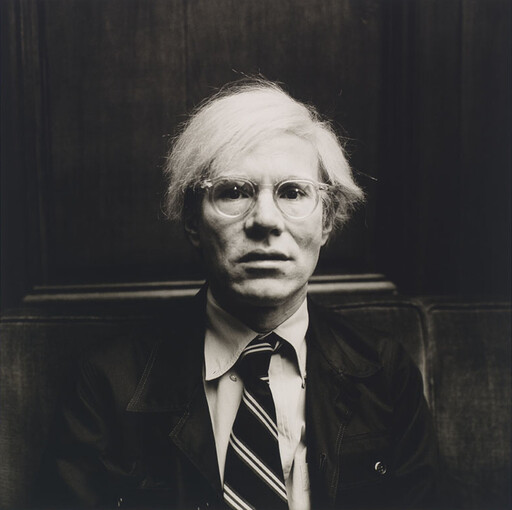 A person with white hair and round glasses wearing a suit and striped tie, sitting still.