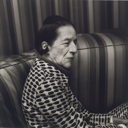 An older woman sits on a striped couch wearing a patterned jacket and looking to the side.
