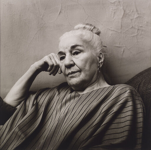 Elderly woman with hair in a bun rests her head on her hand, looking thoughtful.