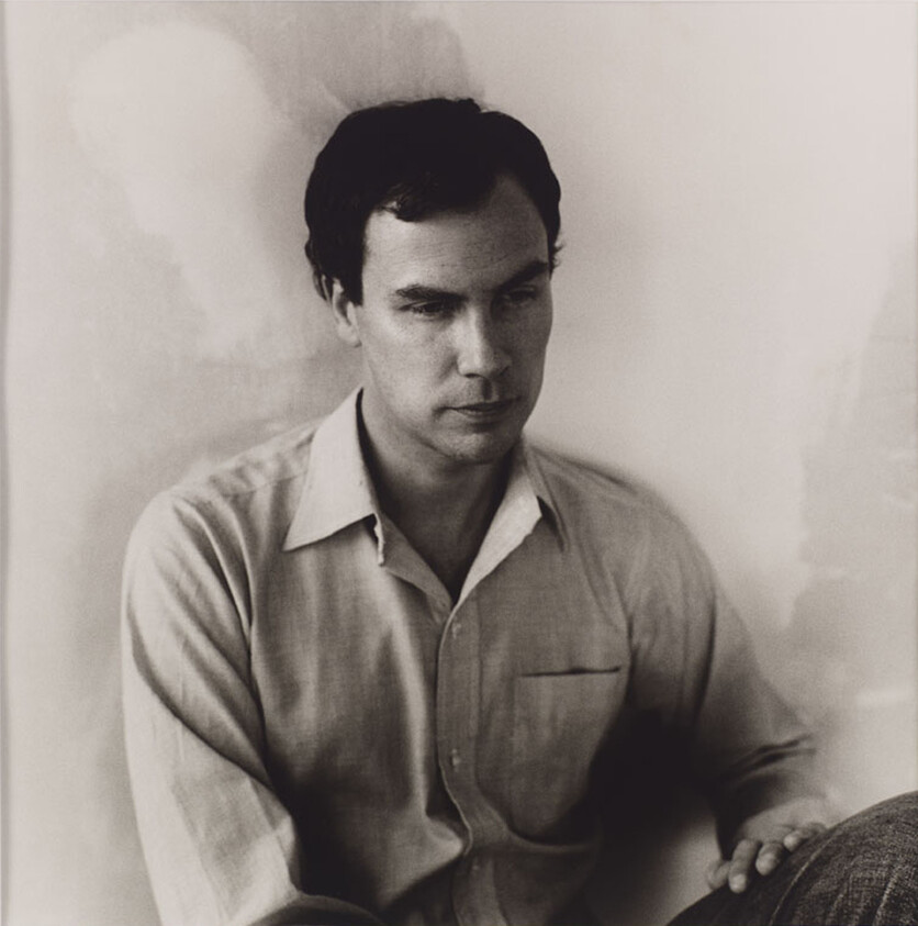 A man in a button-down shirt sits against a wall looking down with a thoughtful expression.