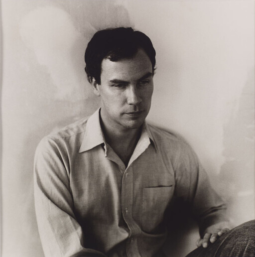 A man in a collared shirt sits against a wall with a thoughtful, serious expression.