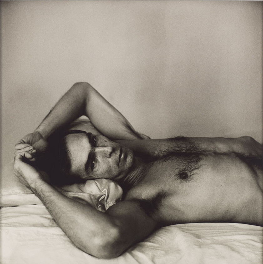 A shirtless man lies on a bed with his arms behind his head, gazing toward the camera.