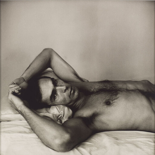 A shirtless man lies on a bed with arms raised behind his head, looking contemplative.