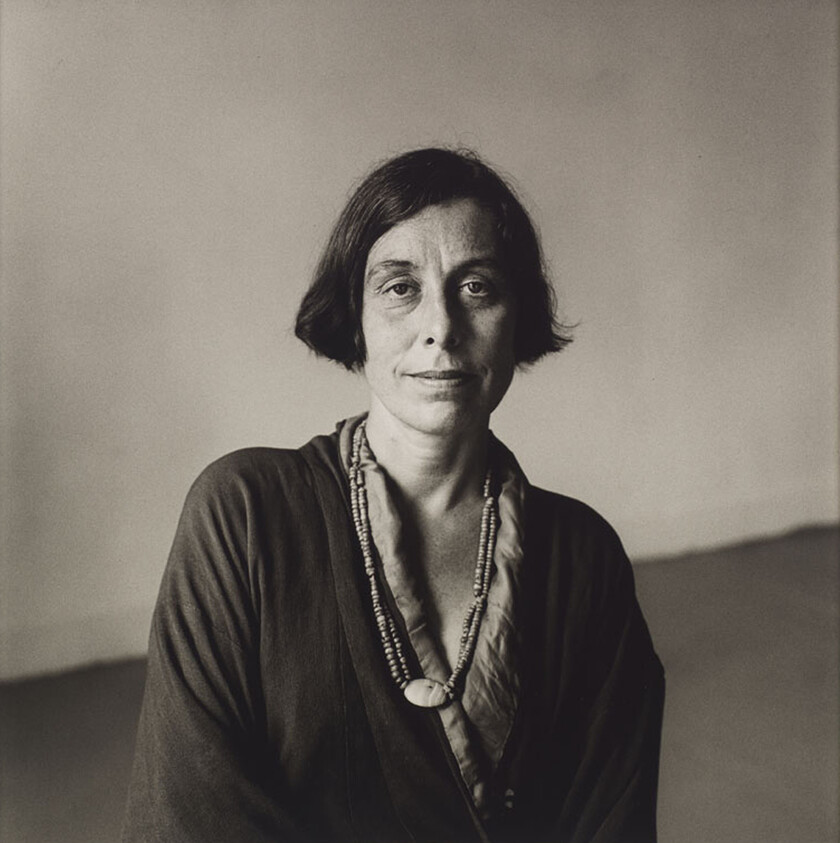 A woman with short hair and a necklace looks directly at the camera, wearing a dark top.