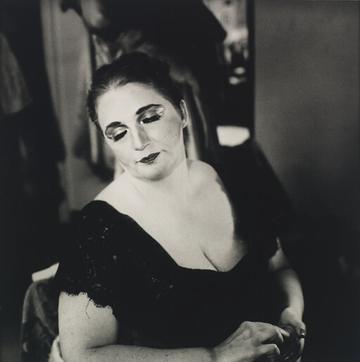 Woman in a black dress with dramatic eye makeup looks down while holding a small object.