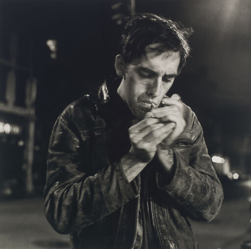 A man in a leather jacket focuses as he lights a cigarette on a dimly lit street.