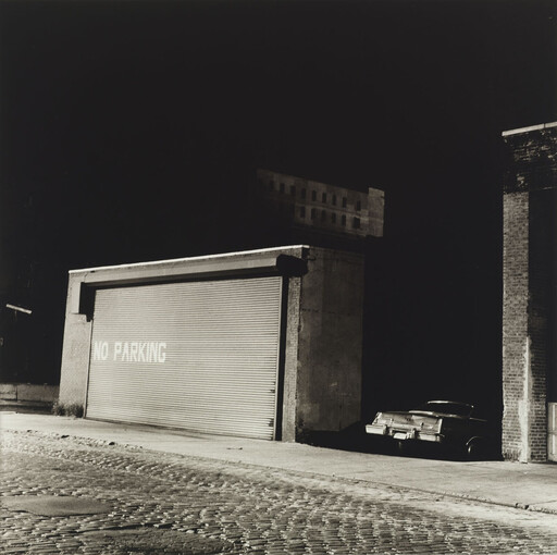 A car is parked in shadow beside a garage door stenciled with the words NO PARKING.