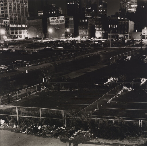 Empty fenced parking lot in foreground with parked cars and illuminated city buildings and a "Park Here" sign.