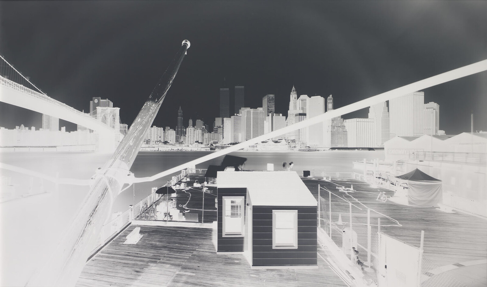 Small shed sits on a wooden pier with bridges and a city skyline across the river.