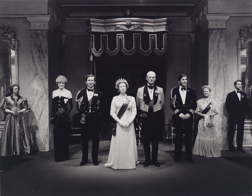 Seven formally dressed figures stand in a grand hall, the central figure wearing a crown.