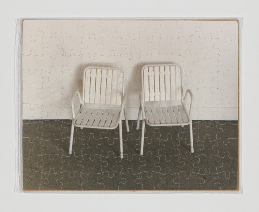 A completed jigsaw puzzle shows two white metal chairs side by side against a wall.