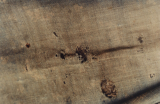 Worn burlap fabric with multiple holes, frayed threads, and a darker stain across the weave.