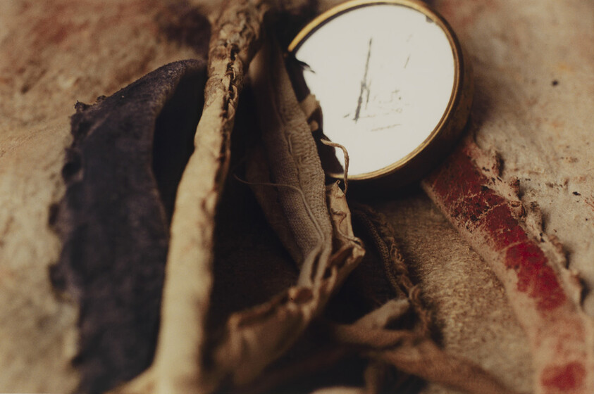 A scratched pocket watch rests among frayed, layered fabric and worn leather straps.