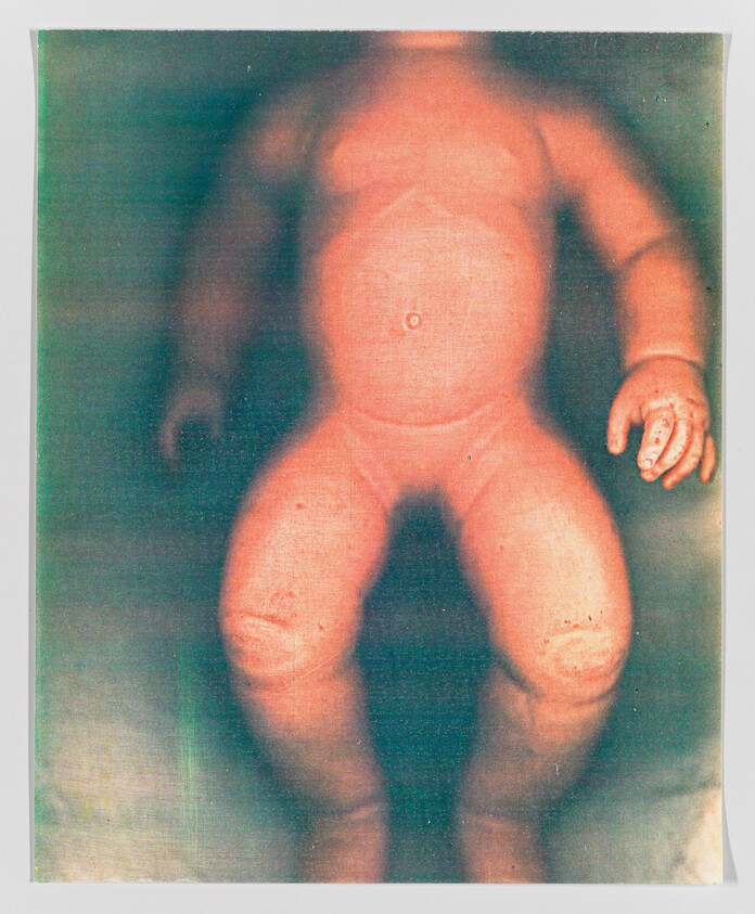 Blurred image of a doll with a distorted, ghostly appearance against a mottled background.