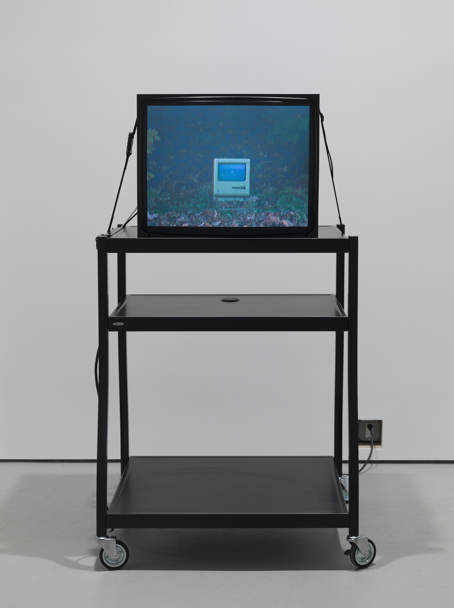 A small CRT monitor on a wheeled metal cart shows a vintage computer icon on screen.