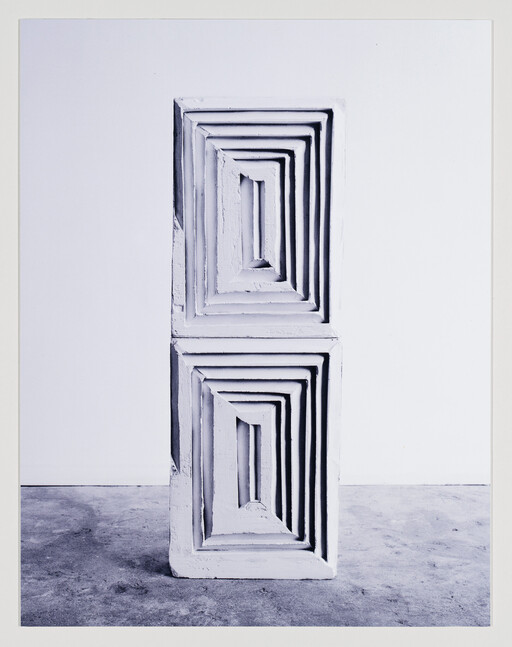 Two stacked carved plaster blocks with concentric recessed rectangles stand on a concrete floor against a white wall.