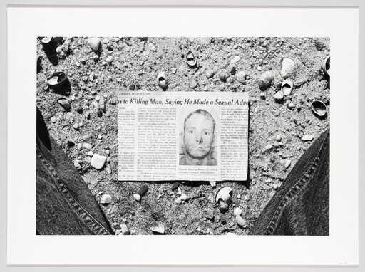 A folded newspaper with a mugshot lies on sandy ground between a person's knees surrounded by shells.