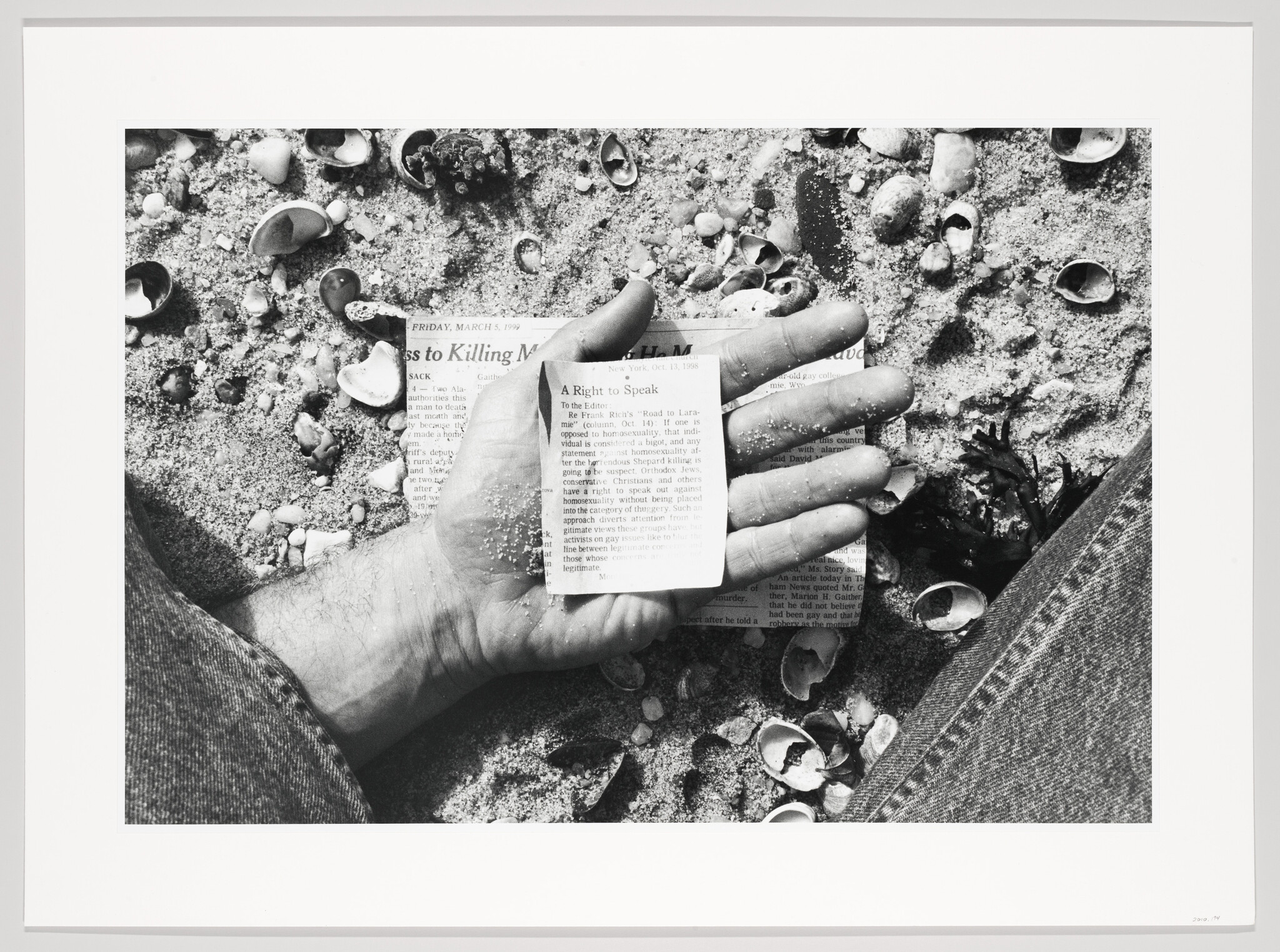 A black and white photo showing a person's hand holding a small, torn piece of newspaper with text visible, resting on a sandy surface scattered with shells. The person is wearing jeans, and only part of their hand and thigh are visible in the frame. The newspaper clipping includes a date, "Friday, March 5," and headlines "A Right to Speak" and "Reasons to Killing M." The image has a border, suggesting it is a print.