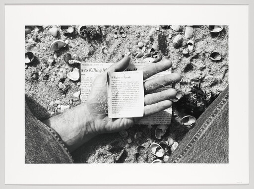 A hand holds a small newspaper clipping on a sandy beach scattered with shells.