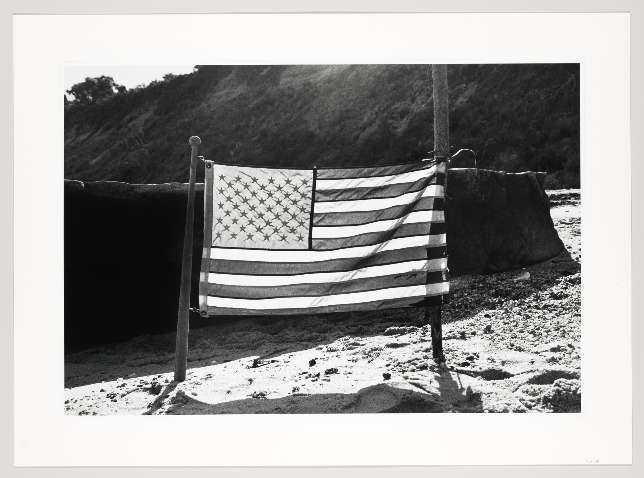 A black and white photograph of an American flag hanging on a pole, with a sandy beach in the foreground and a hill with vegetation in the background.