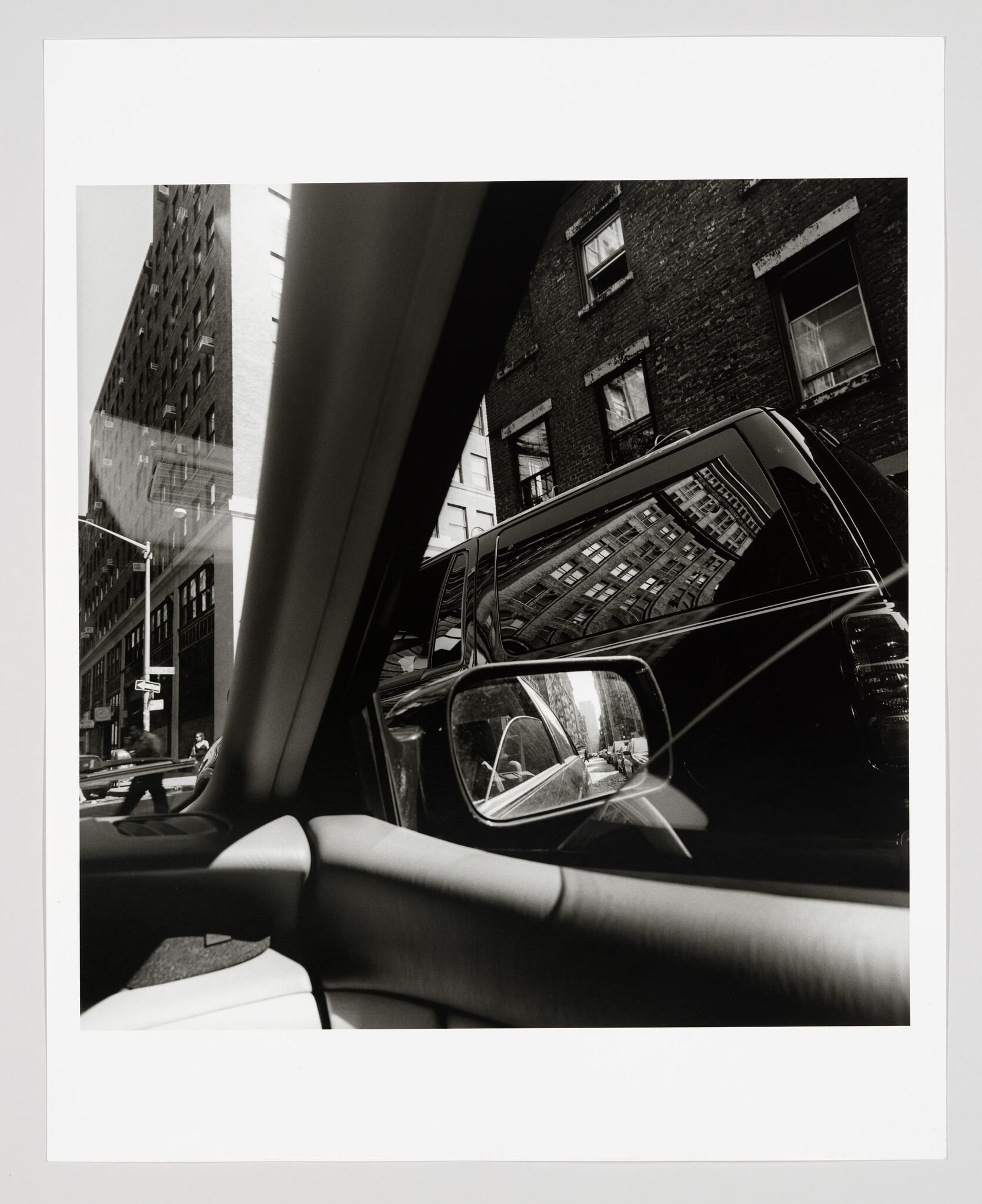 Car interior shows side mirror reflecting tall city buildings and a black SUV parked beside.