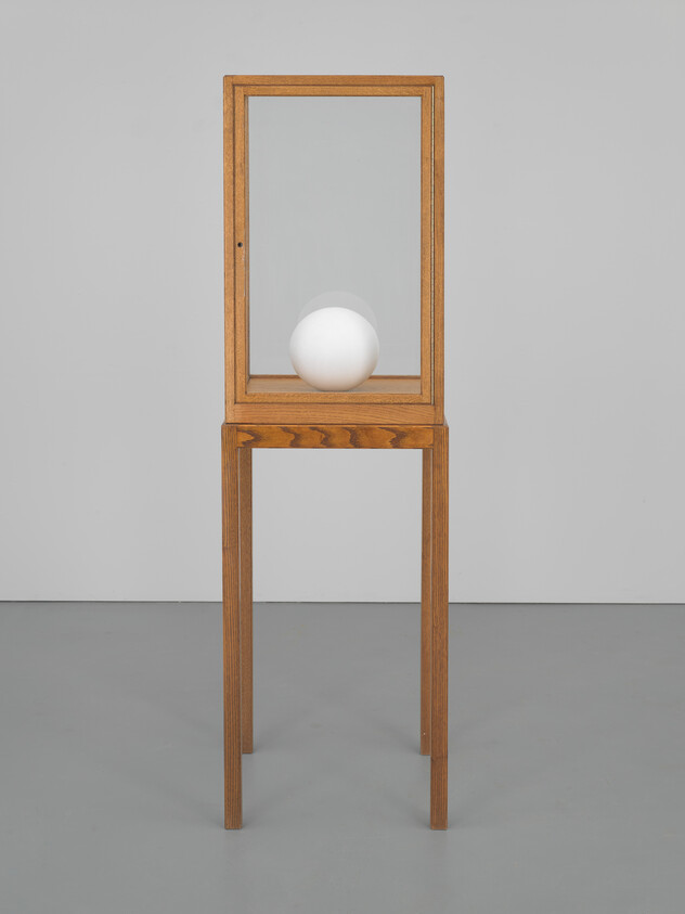 A wooden glass display cabinet on tall legs contains a single white sphere.