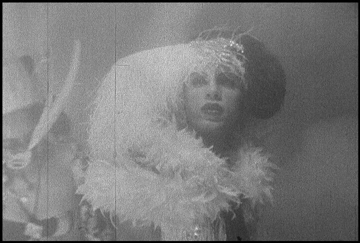 A person wearing an elaborate feathered hat and boa looks upward with a surprised expression.