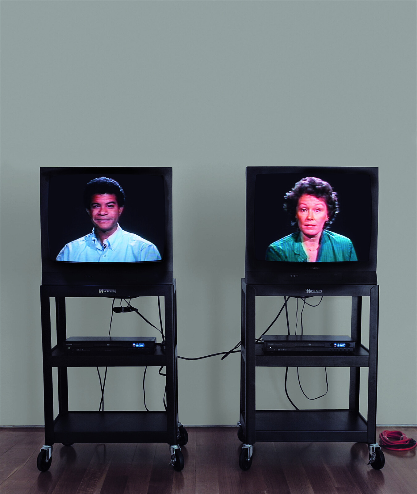 Two CRT televisions on wheeled carts display a man smiling and a woman speaking.