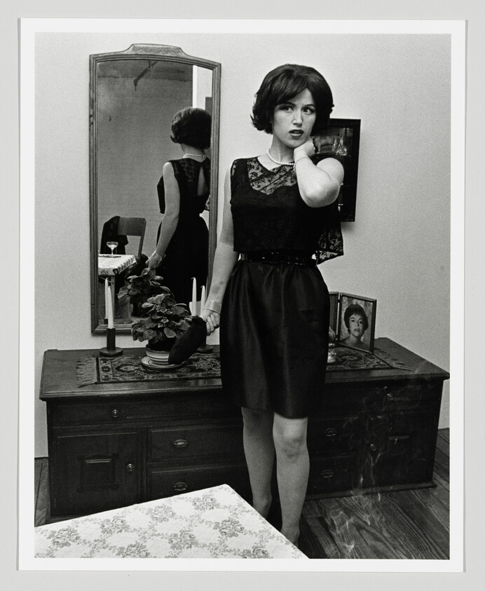 A woman in a black dress stands by a dresser, touching her neck, with her reflection in the mirror.