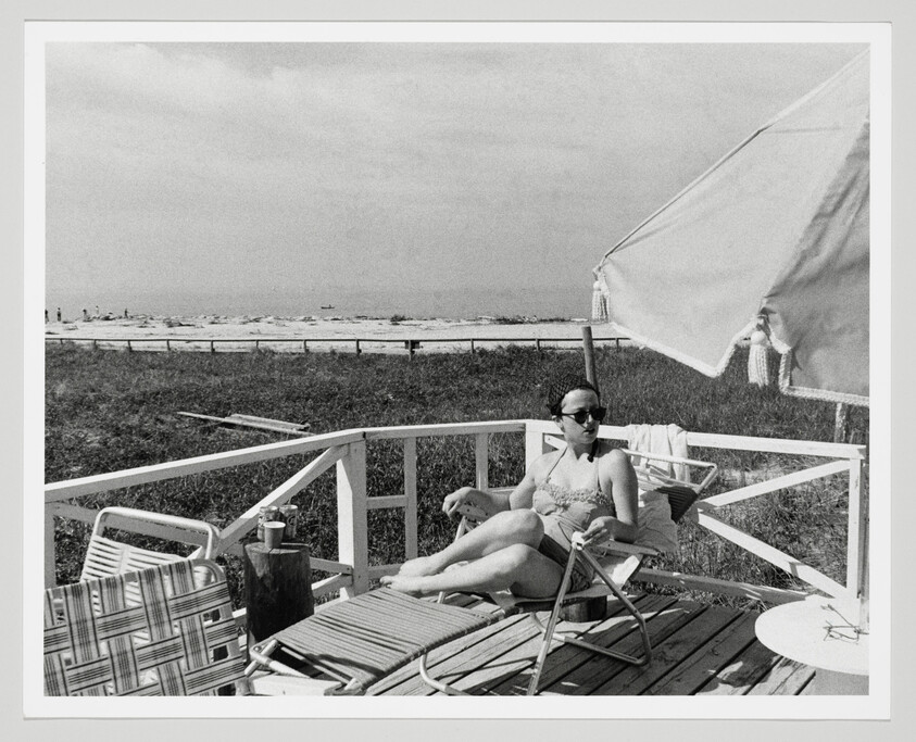 A woman in sunglasses relaxes on a deck chair under a large beach umbrella near the shore.
