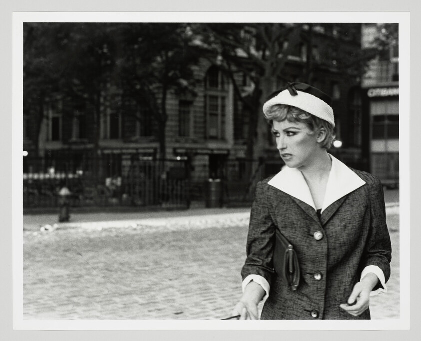 A woman in a vintage suit and hat walks along a cobblestone street, looking to her right.
