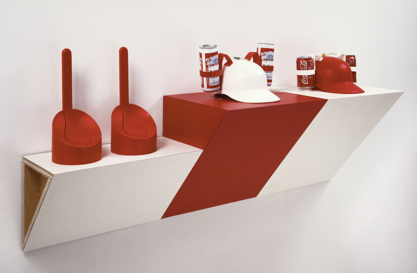 A wall-mounted red and white shelf displaying two red toilet scoops and two helmets with canned drinks attached.