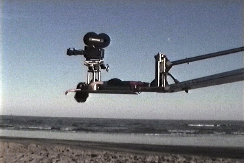 A film camera on a long jib arm filming over a sandy beach and ocean.