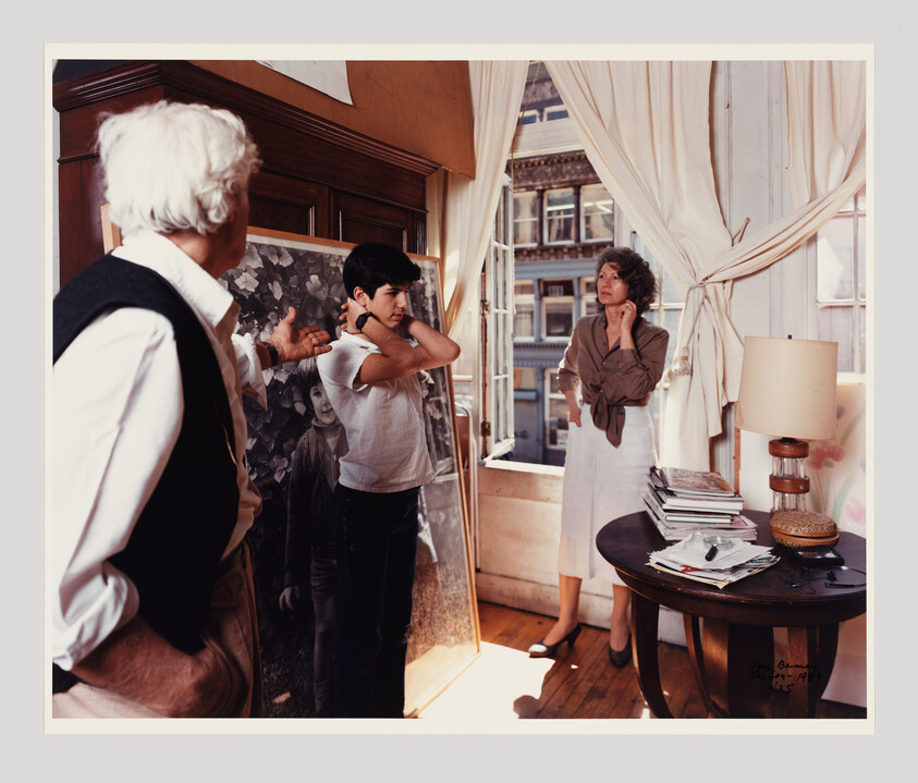 An older man gestures at a teenage boy adjusting his shirt while a woman watches by the window.