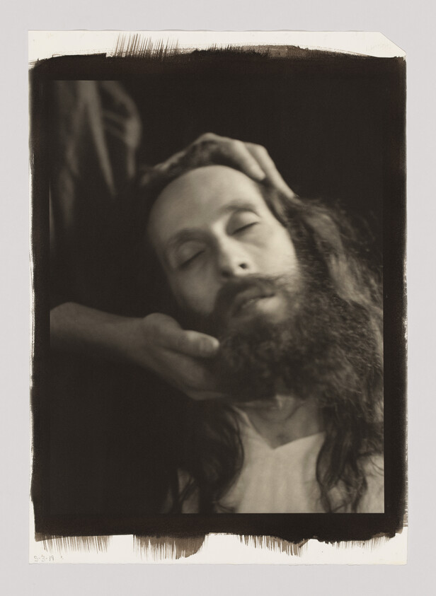 A bearded man with closed eyes has his head gently cradled by another person's hands.