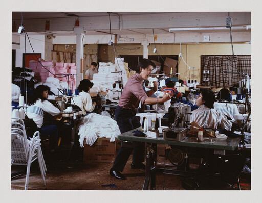 Factory workers sew garments while a manager gestures and shows fabric to a seated woman.