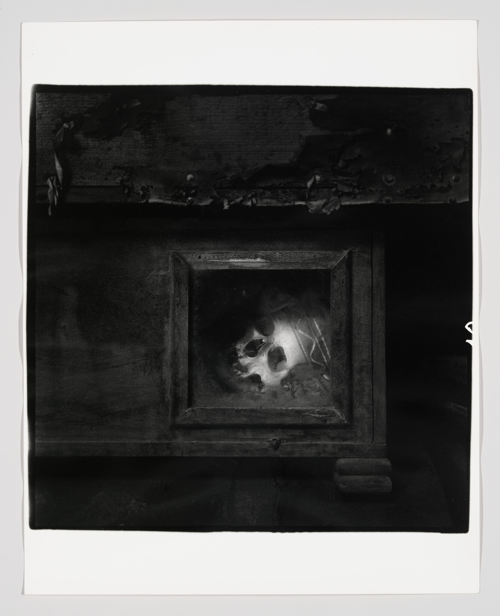 A human skull lies inside a small framed wooden panel on a dark, aged surface.