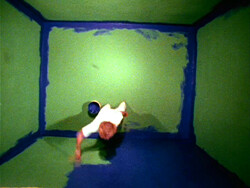 Person painting the floor and lower walls of a green room with blue paint, seen from above.