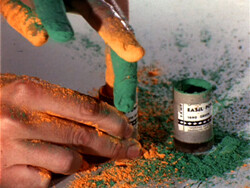 Hands covered in green and orange powder touch small containers, with powder scattered on the surface.