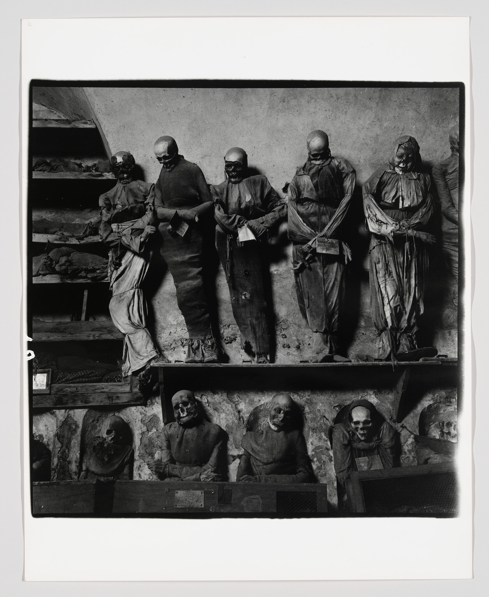 Several mummified corpses dressed in old clothing stand and sit displayed in a dim crypt.