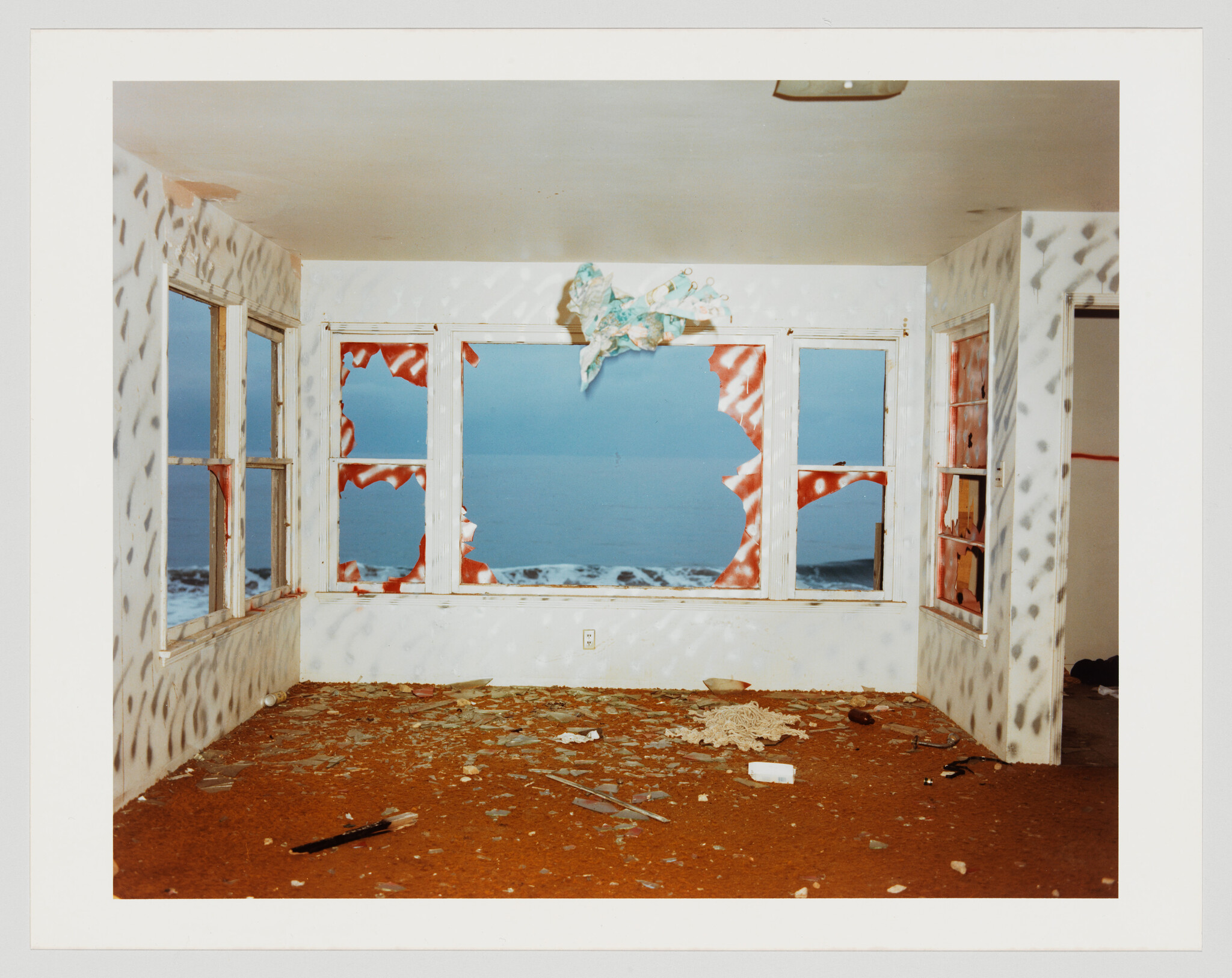 Abandoned room with shattered windows facing the ocean and debris scattered across the floor.
