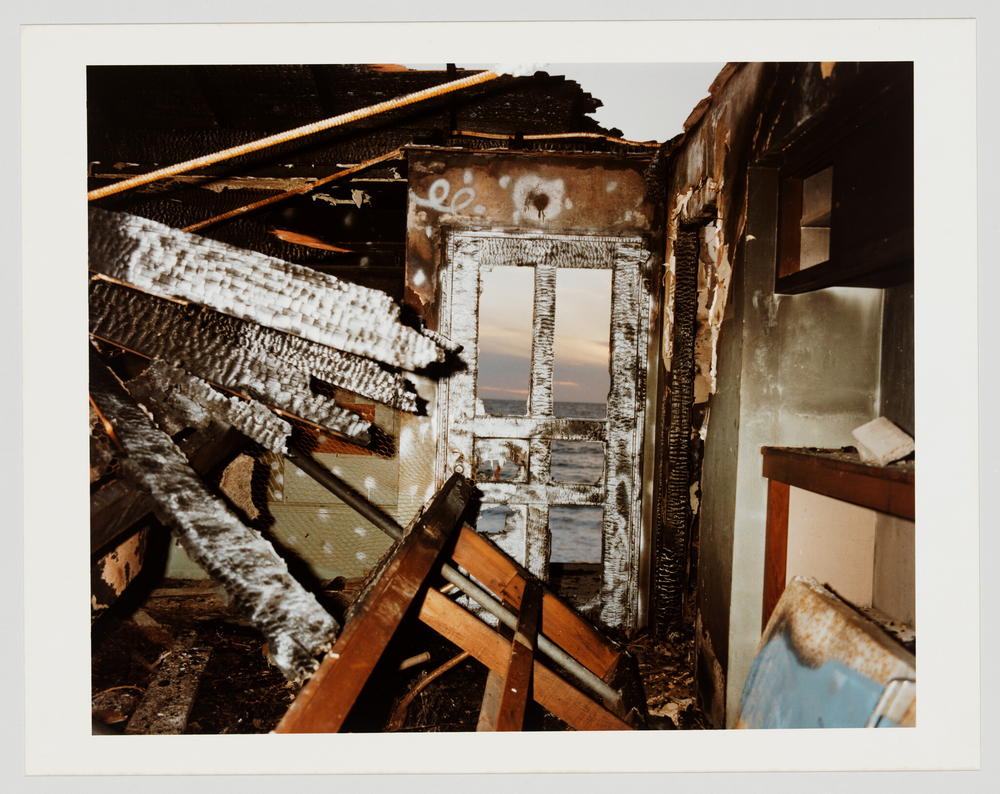 Charred interior of a burned room with a damaged door opening to a calm ocean at sunset.