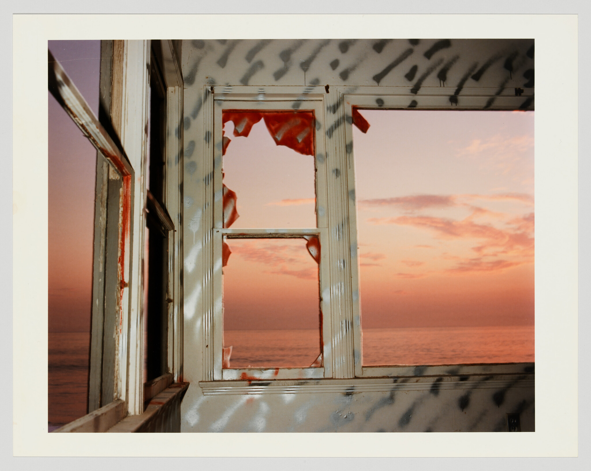 Torn red curtains hang in a weathered window framing a pink sunset over the ocean.
