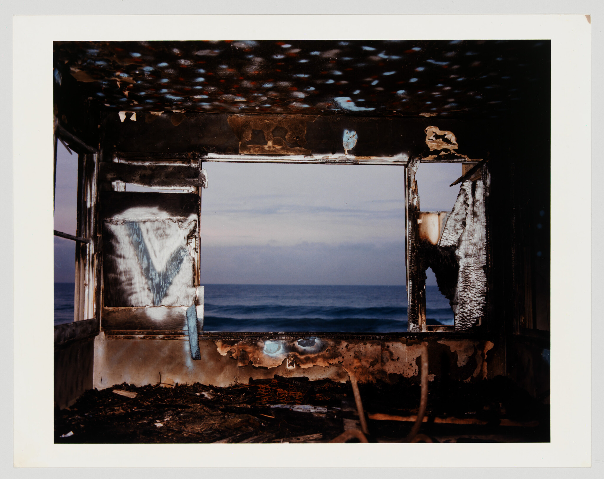 Burned room interior with charred walls framing a calm ocean view through a large window.