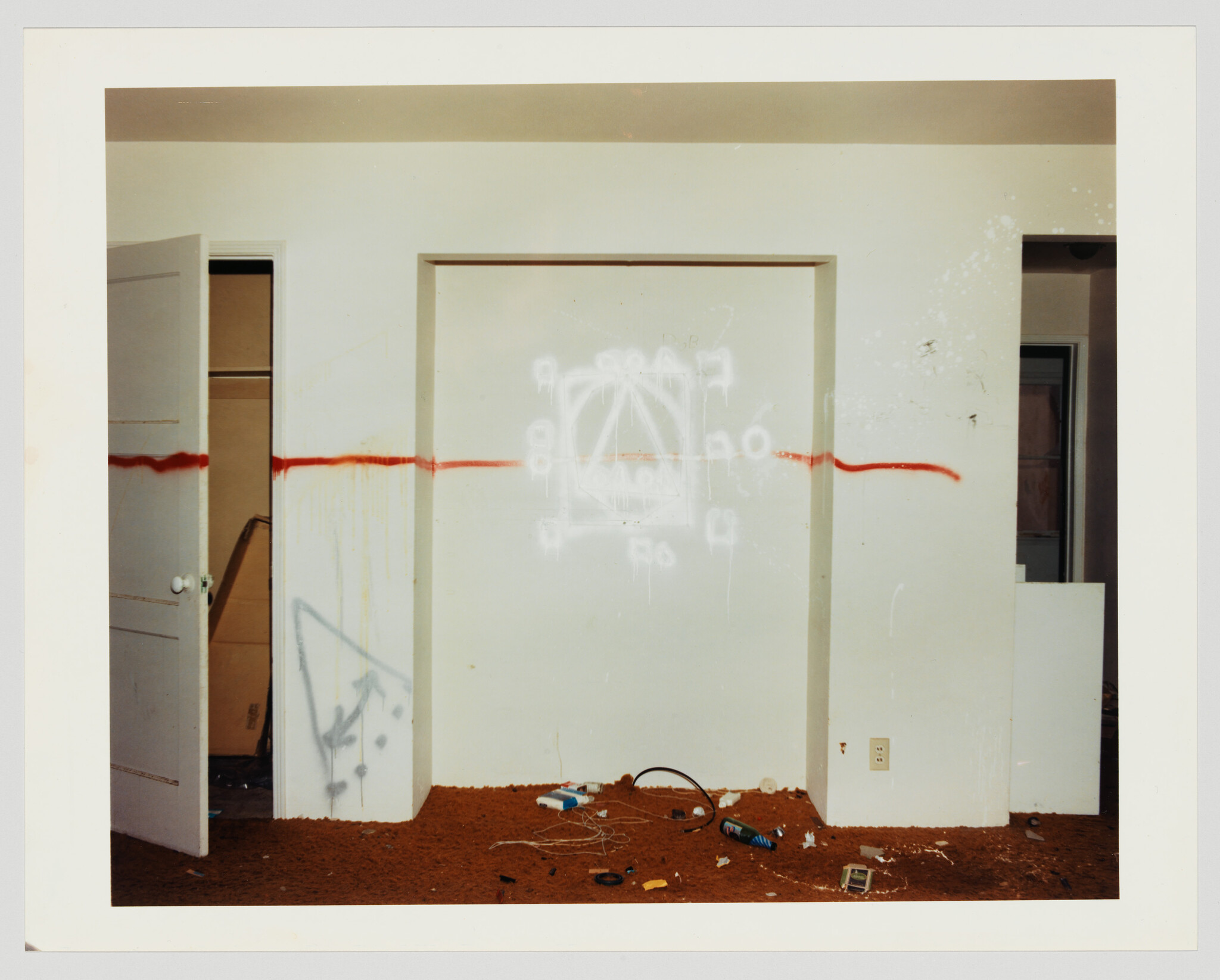Empty room with a red spray-painted line and white graffiti above a cluttered floor.