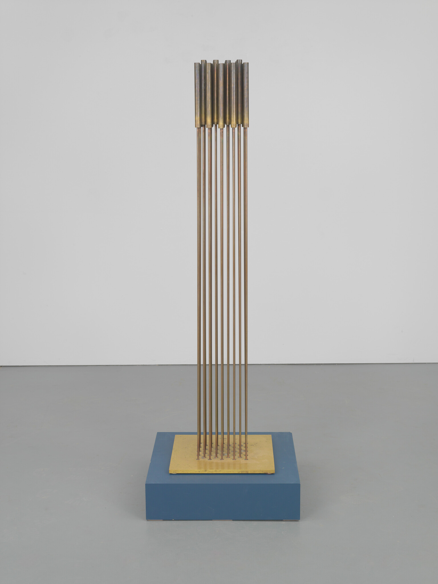 Tall sculpture of thin vertical brass rods mounted on a square yellow plate atop a blue base.
