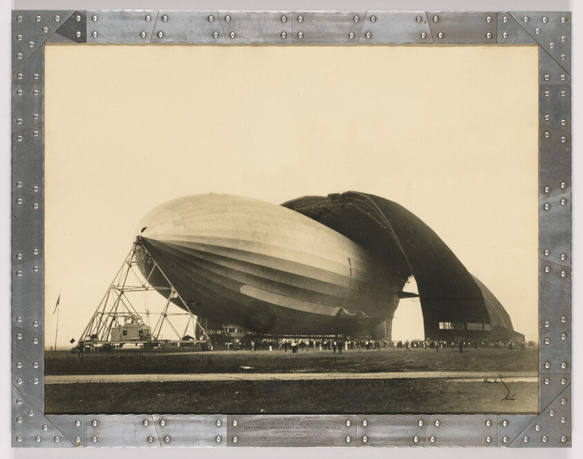 Large airship being assembled near a curved hangar while workers and spectators stand nearby.